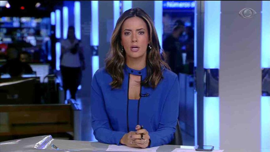 Paloma Tocci veste uma blusa azul sentada na bancada do Jornal da Band