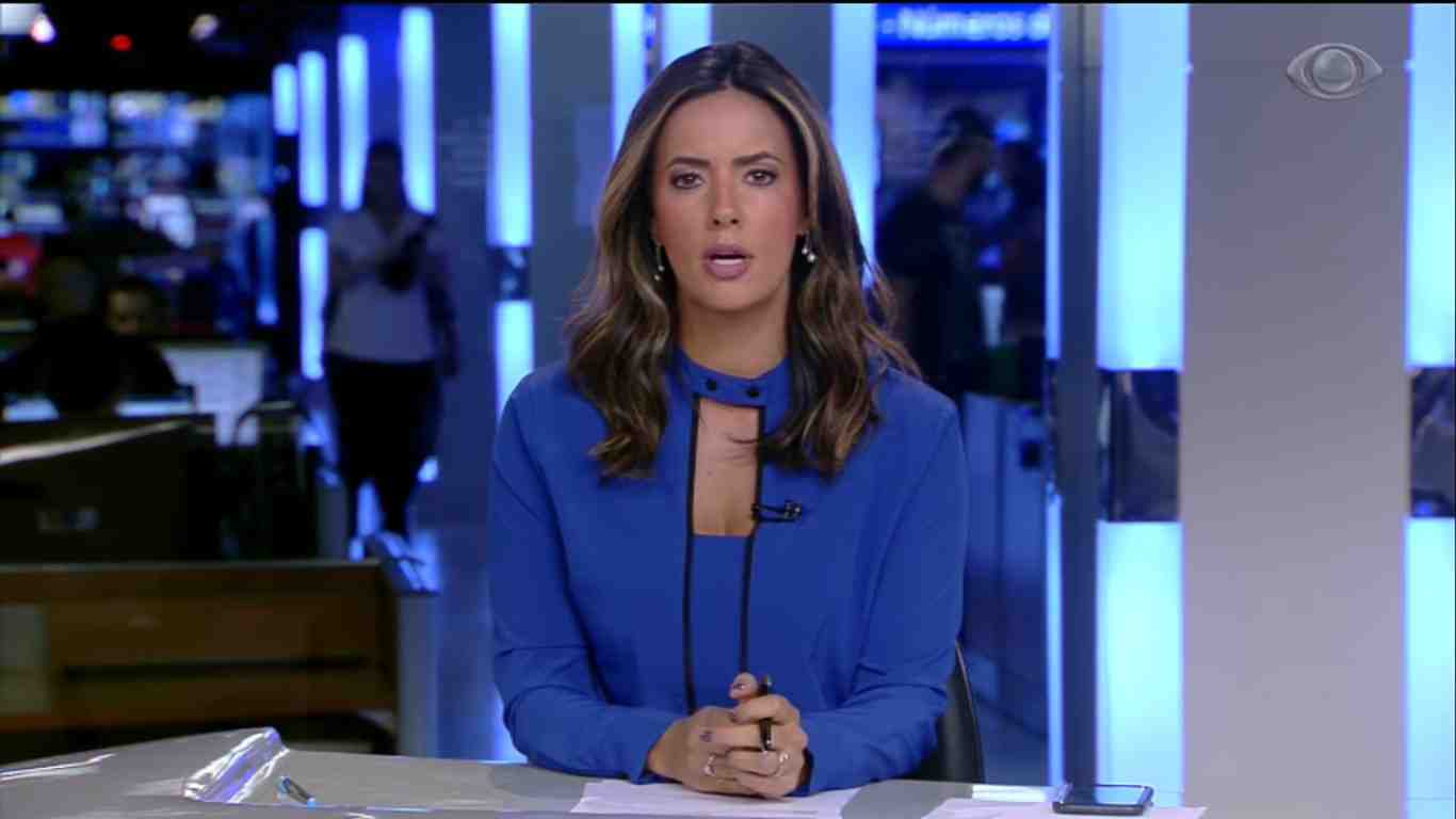 Paloma Tocci veste uma blusa azul sentada na bancada do Jornal da Band