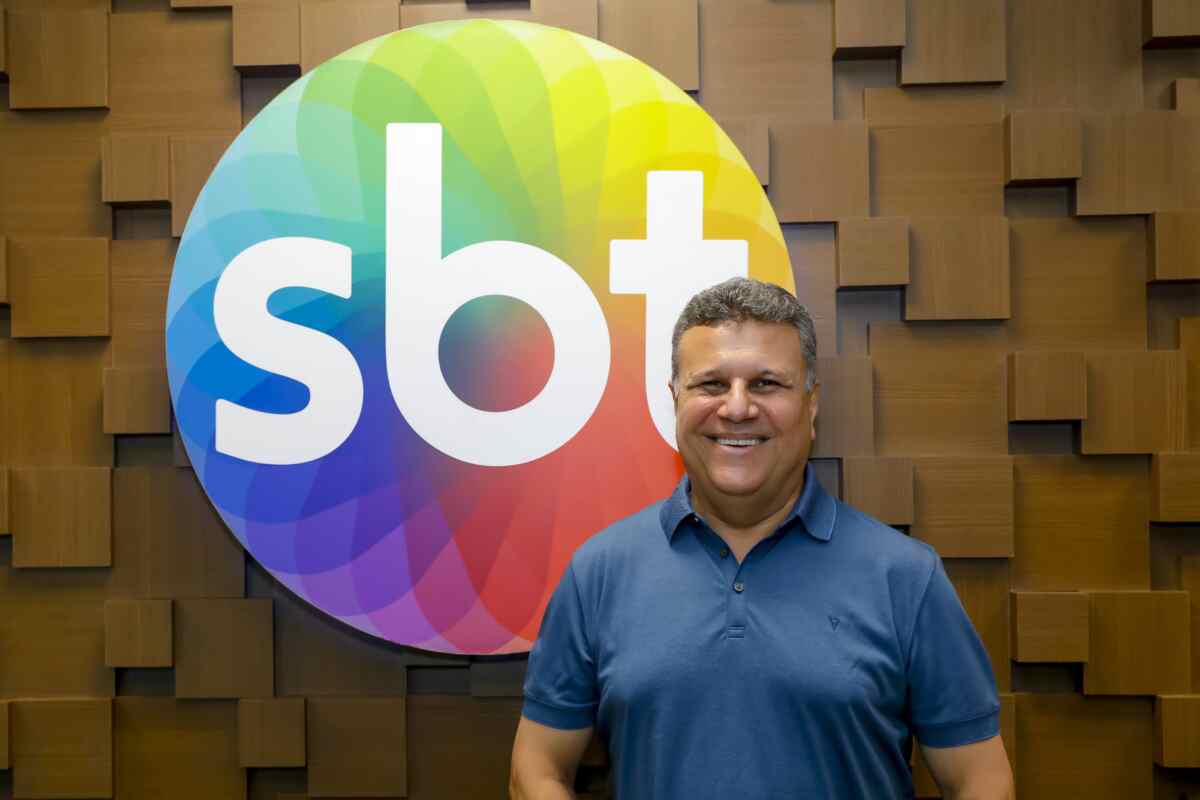 Narrador Téo José em frente a uma banner de parede com o logo do SBT