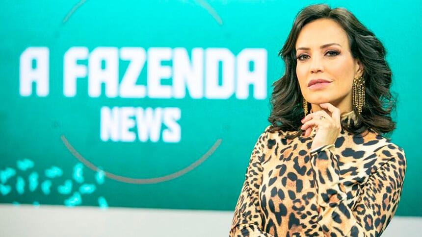 Fabiana Oliveira com a mão no queixo, posando para foto no cenário do programa A Fazenda News