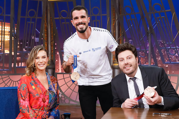 Michelle Lenhardt (sentada) ao lado de Bruno Fratus (em pé e segurando uma medalha) e Danilo Gentili (sentado e segurando uma medalha) nos estúdios do The Noite no SBT