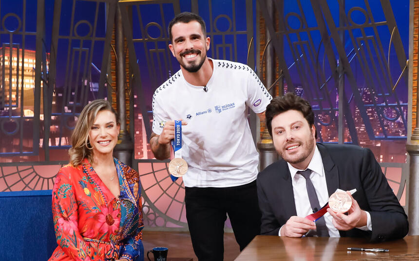 Michelle Lenhardt (sentada) ao lado de Bruno Fratus (em pé e segurando uma medalha) e Danilo Gentili (sentado e segurando uma medalha) nos estúdios do The Noite no SBT