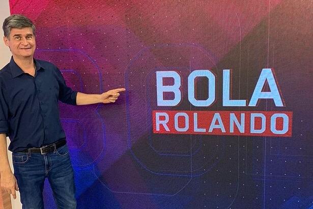 Fábio Piperno nos estúdios do Bola Rolando, no BandSports, em pé, apontando para o logo do programa que está ao seu lado