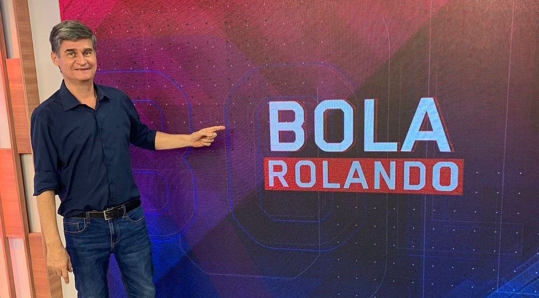 Fábio Piperno nos estúdios do Bola Rolando, no BandSports, em pé, apontando para o logo do programa que está ao seu lado