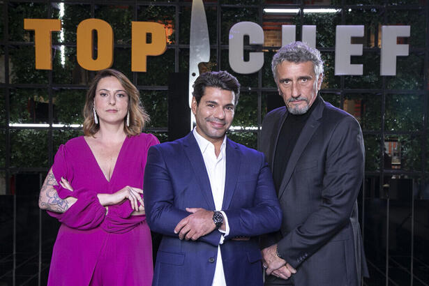 Ailin Aleixo, Felipe Bronze e Emmanuel Bassoleil em uma foto de divulgação do Top Chef Brasil, à frente de um banner com o logo do reality