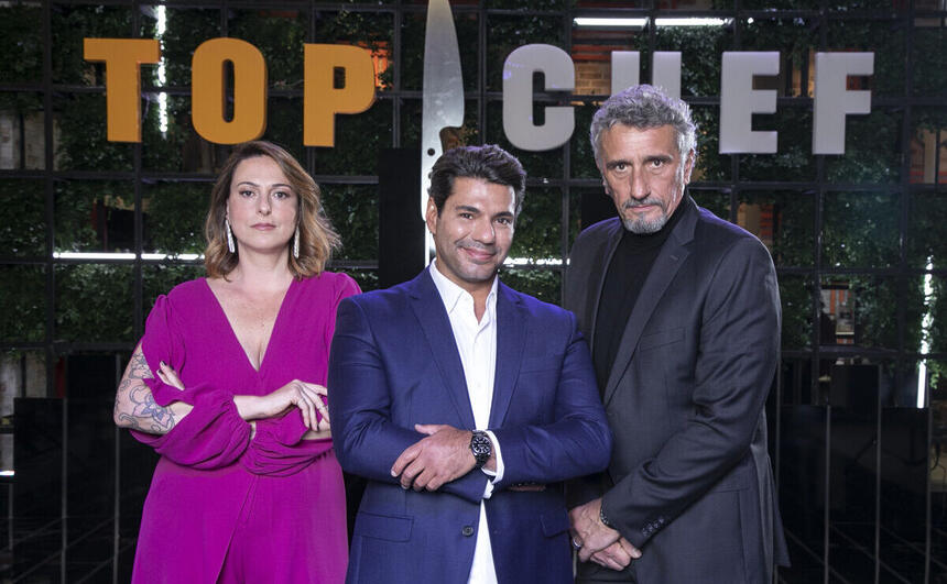 Ailin Aleixo, Felipe Bronze e Emmanuel Bassoleil em uma foto de divulgação do Top Chef Brasil, à frente de um banner com o logo do reality