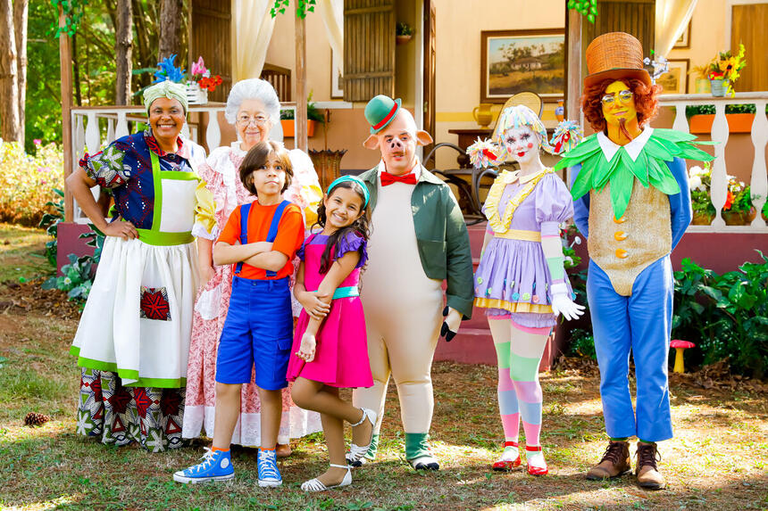 Atores caracterizados com os personagens do sítio posam em frente ao cenário do Picapau Amarelo