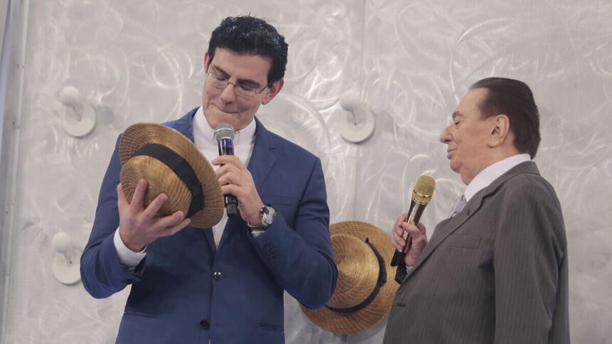 Padre Reginaldo Manzotti segurando um chapéu ao lado de Raul Gil, nos estúdios do programa