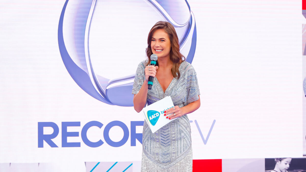 Thaís Pacholek sorrir enquanto apresenta o Teleton 2021. Por trás da atriz o logo da Record TV, sua nova casa, aparece no telão,