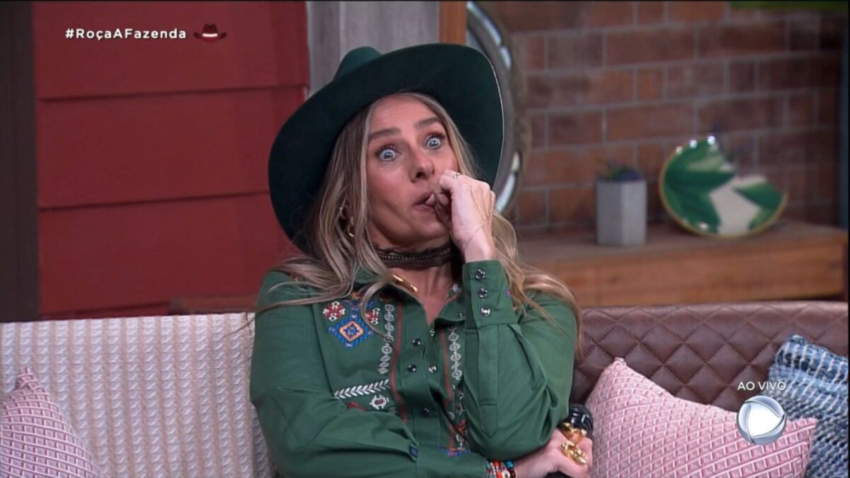 Adriane Galisteu, assustada e sentada no cenário de A Fazenda, com o dedo na boca e acompanhando o barraco na formação da roça