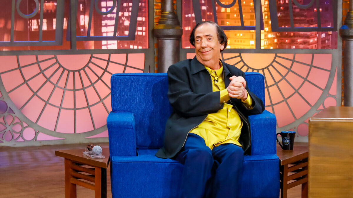 Marcos Oliveira sentado em uma poltrona azul nos estúdios do The Noite no SBT