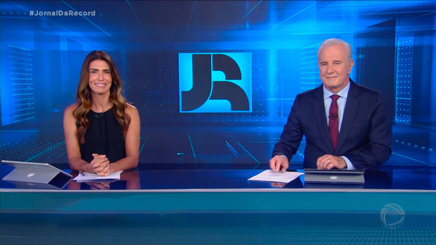 Janine Borba ao lado de Celo Freitas, ambos estão sorrindo, após se despedirem do público no final do Jornal da Record desta terça-feira, 28 de dezembro de 2021