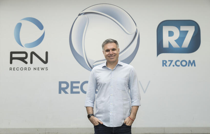 Narrador Marco de Vargas posando para foto em frente aos logos da Record TV, Record News e R7