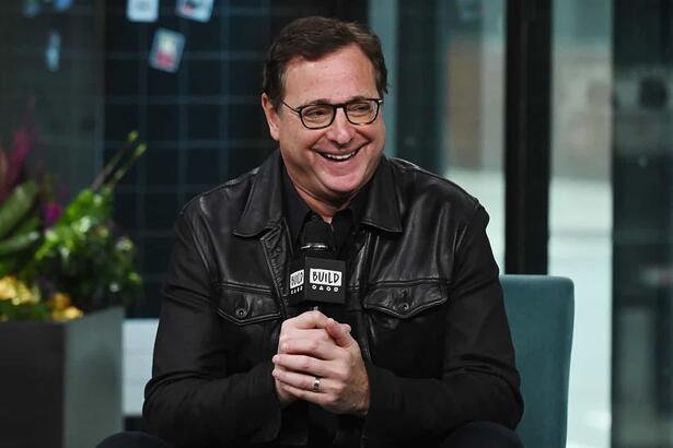 Bob Saget durante participação em um programa, com um microfone na mão