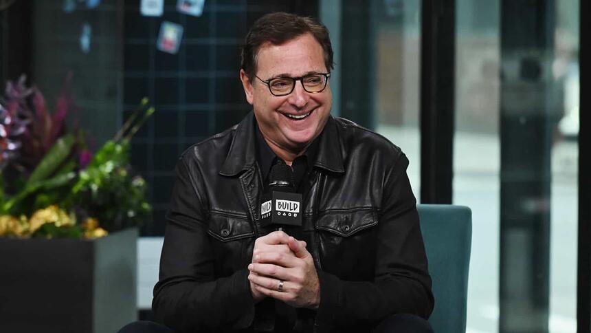 Bob Saget durante participação em um programa, com um microfone na mão