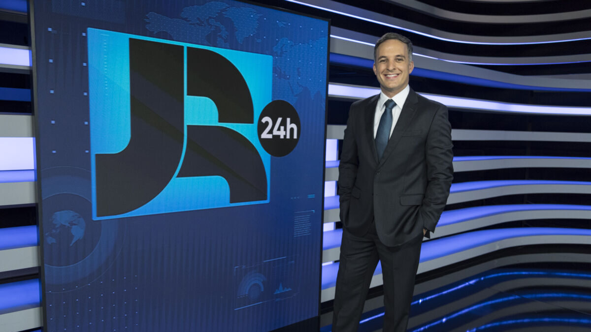 Leandro Stoliar de terno, parando em frente ao telão do JR 24h no estúdio e com as mãos nos bolsos