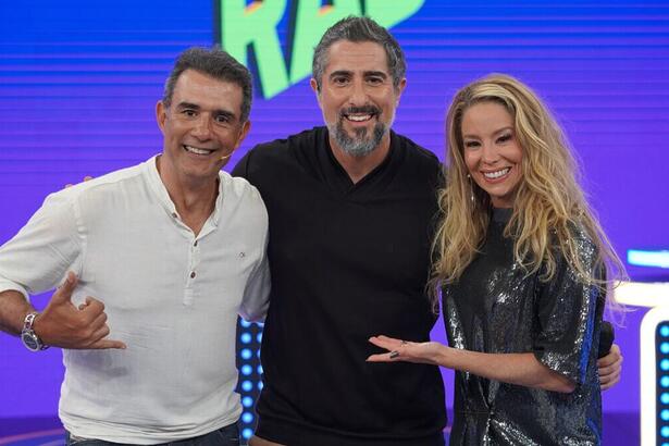 Marcos Pasquim, de blusa branca, abraçado com Marcos Mion, de blusa preta, e Danielle Winits, de vestido cinza brilhoso, nos estúdios do Caldeirão