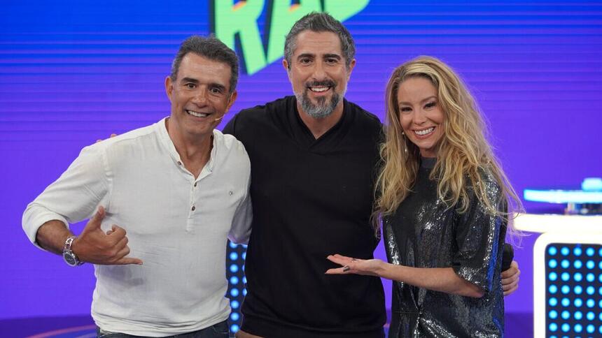 Marcos Pasquim, de blusa branca, abraçado com Marcos Mion, de blusa preta, e Danielle Winits, de vestido cinza brilhoso, nos estúdios do Caldeirão