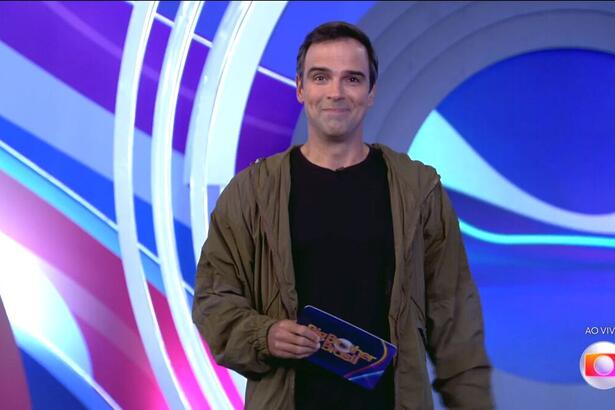 Tadeu Schmidt com uma blusa preto, um casaco marrom por cima e uma ficha na mão, no cenário do BBB 22 durante o episódio de estreia