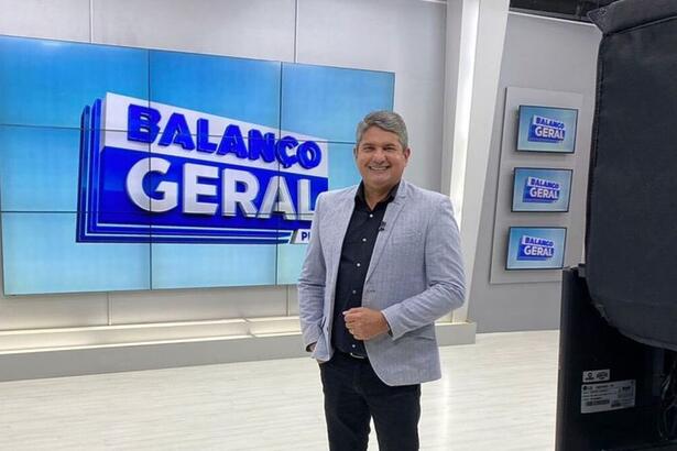 Gernand Lopes de terno cinza. Ele está sorrindo, em frente ao telão do Balanço Geral PE