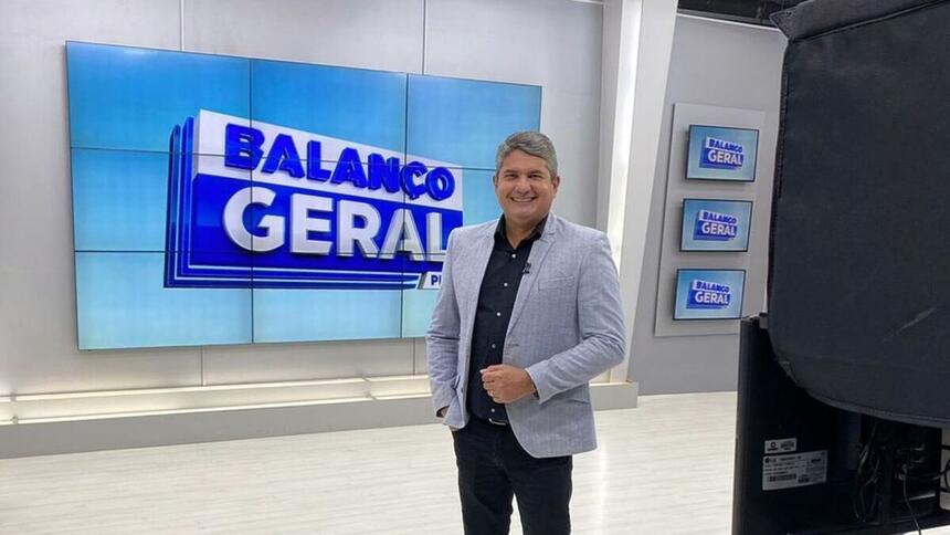 Gernand Lopes de terno cinza. Ele está sorrindo, em frente ao telão do Balanço Geral PE