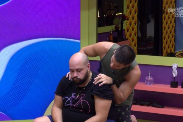 Tiago Abravanel sentado na sala do BBB, com Arthur Aguiar em pé, o abraçando e o consolando por trás