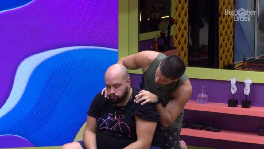 Tiago Abravanel sentado na sala do BBB, com Arthur Aguiar em pé, o abraçando e o consolando por trás