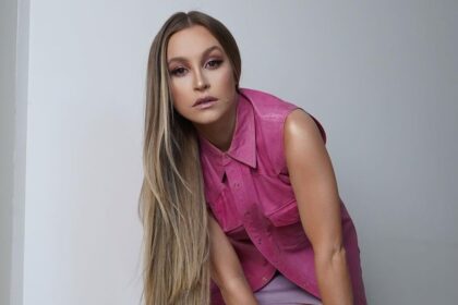 Carla Diaz, em uma foto posada, utilizando uma camisa rosa e com o corpo reclinado para frente