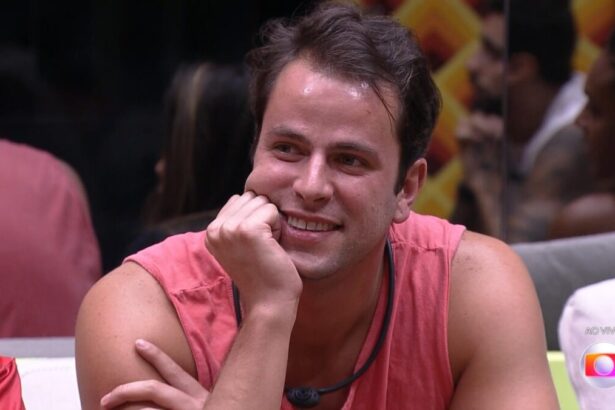 Gustavo com a mão no queixo sentado no sofá da sala do BBB 22 recebe a notícia que está no PAredão
