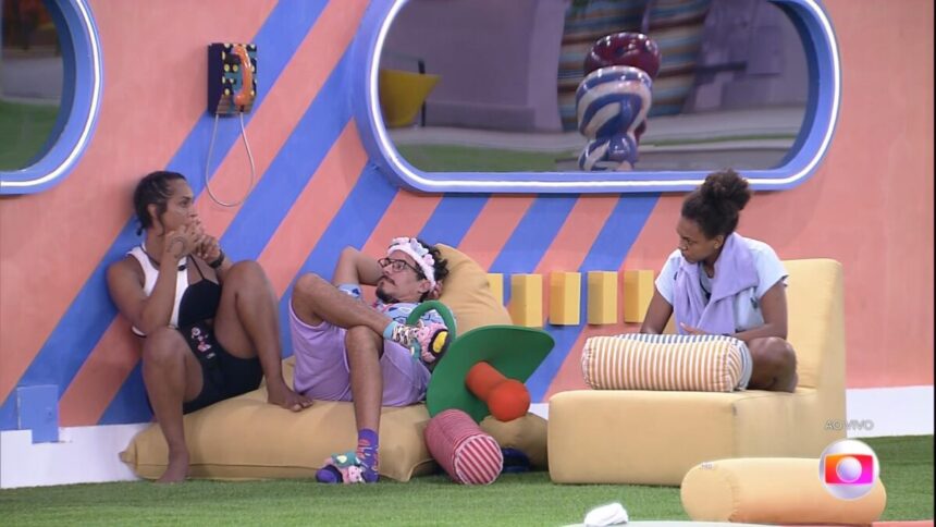 Linn da Quebrada sentada abaixo do Big Fone, ao lado dela Elizer e Jessilan sentada no sofá do BBB 22