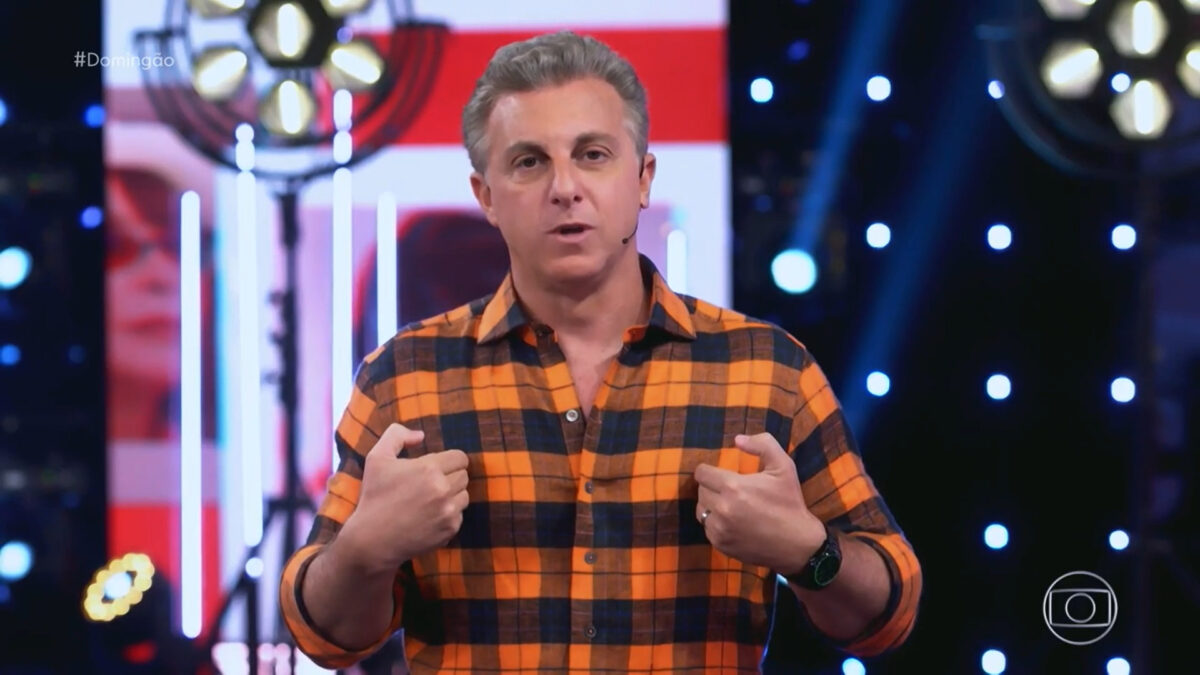Luciano Huck, com uma camisa xadrez, apontando os dedos para si no estúdio do Domingão