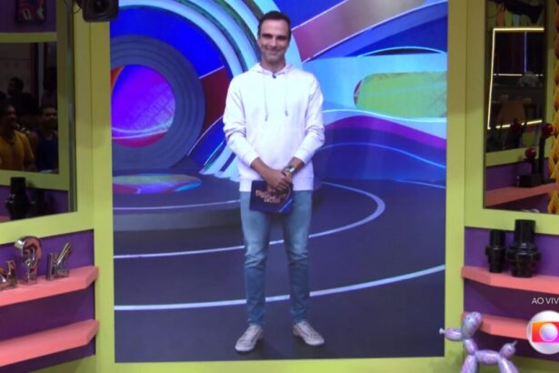 Tadeu aparecendo para falar com participantes do BBB 22 através do televisor. Tadeu veste camisa de manga longa branca e calça comprida.