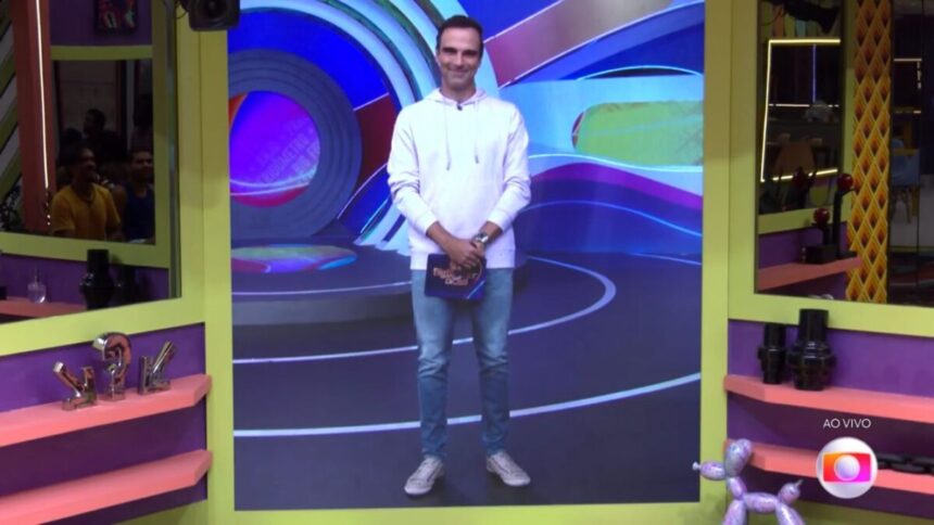 Tadeu aparecendo para falar com participantes do BBB 22 através do televisor. Tadeu veste camisa de manga longa branca e calça comprida.