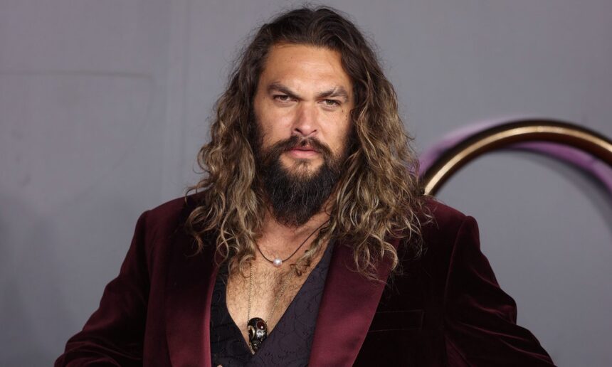 Jason Momoa vestido com um terno vinho e cabelo solto