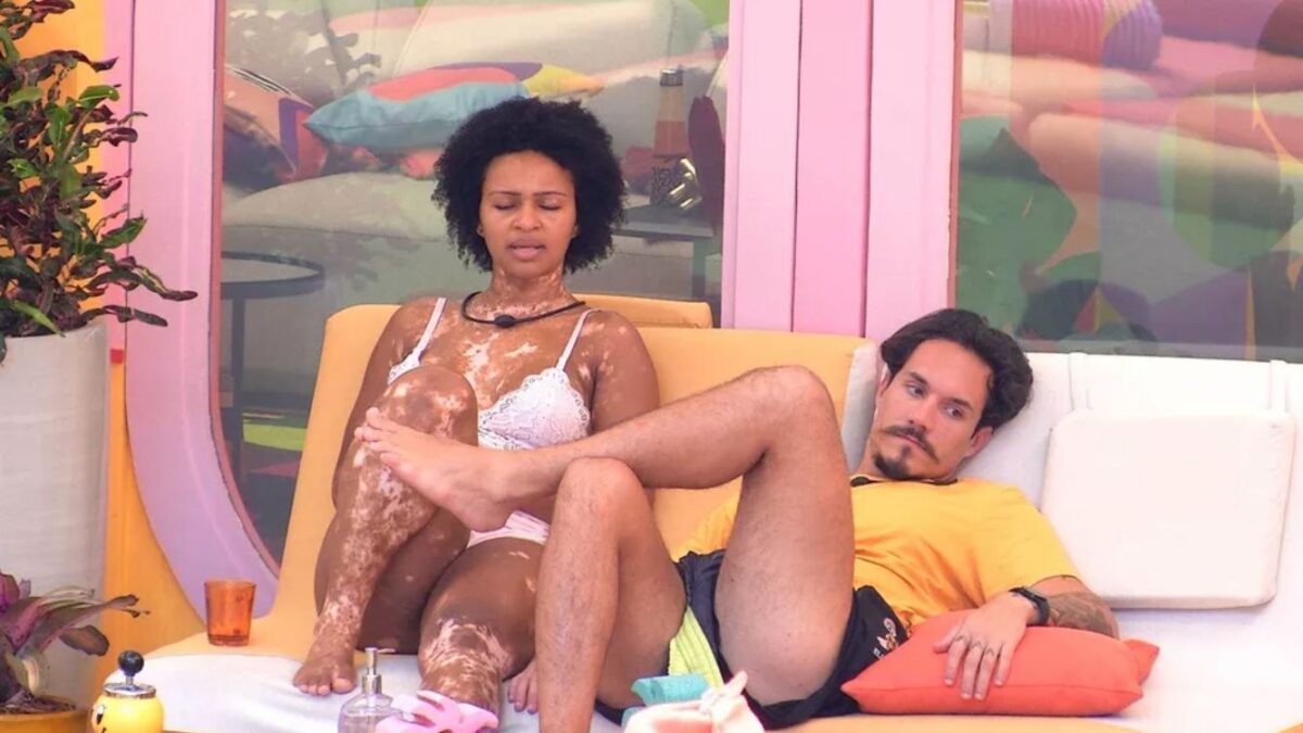 Natália de blusa branca sentado ao lado de Eliezer de pernas cruzadas.