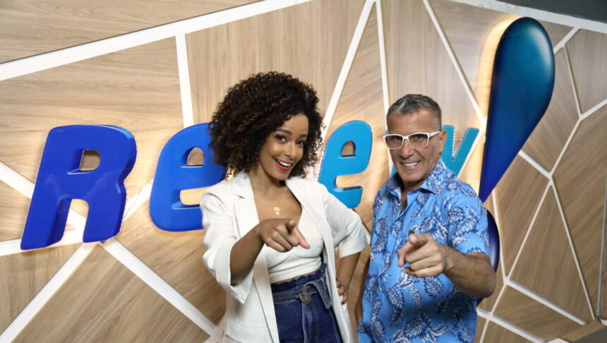 Alinne Prado e Eri Johnson, abraçados em frente ao telão com o logo da RedeTV! e apontando com o dedo para a câmera