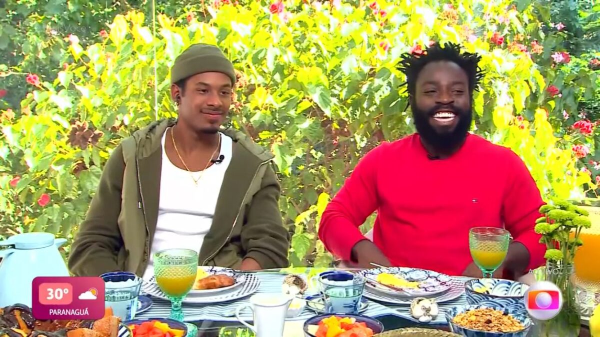 Paulo André e Douglas sentados com uma mesa posta de café da manhã.