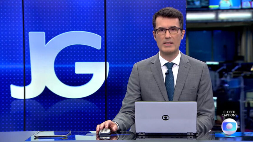 Fabio Turci sentado na bancada do Jornal da Globo, com o telão ao fundo e um notebook na bancada