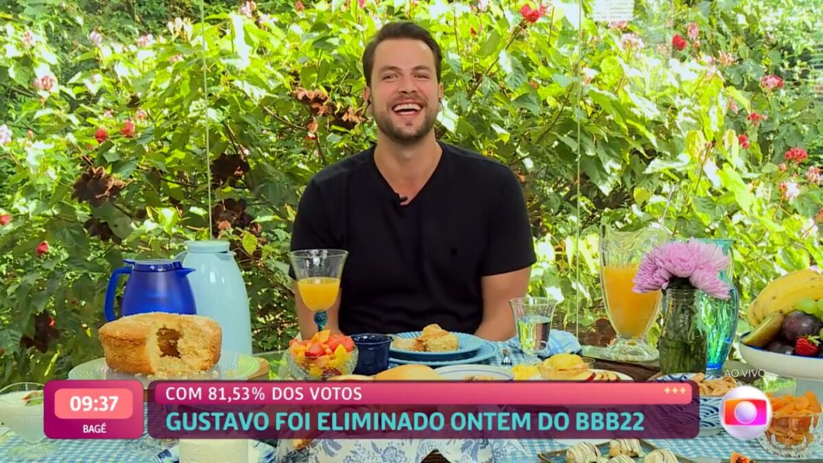 Gustavo sorrindo sentado de frente para uma mesa de café da manhã no Mais Você.