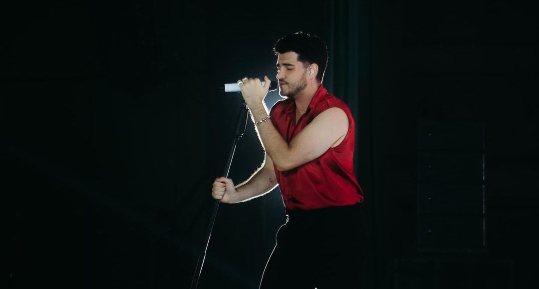 João vestindo uma blusa vermelha e um microfone com tripé na mão, durante um show