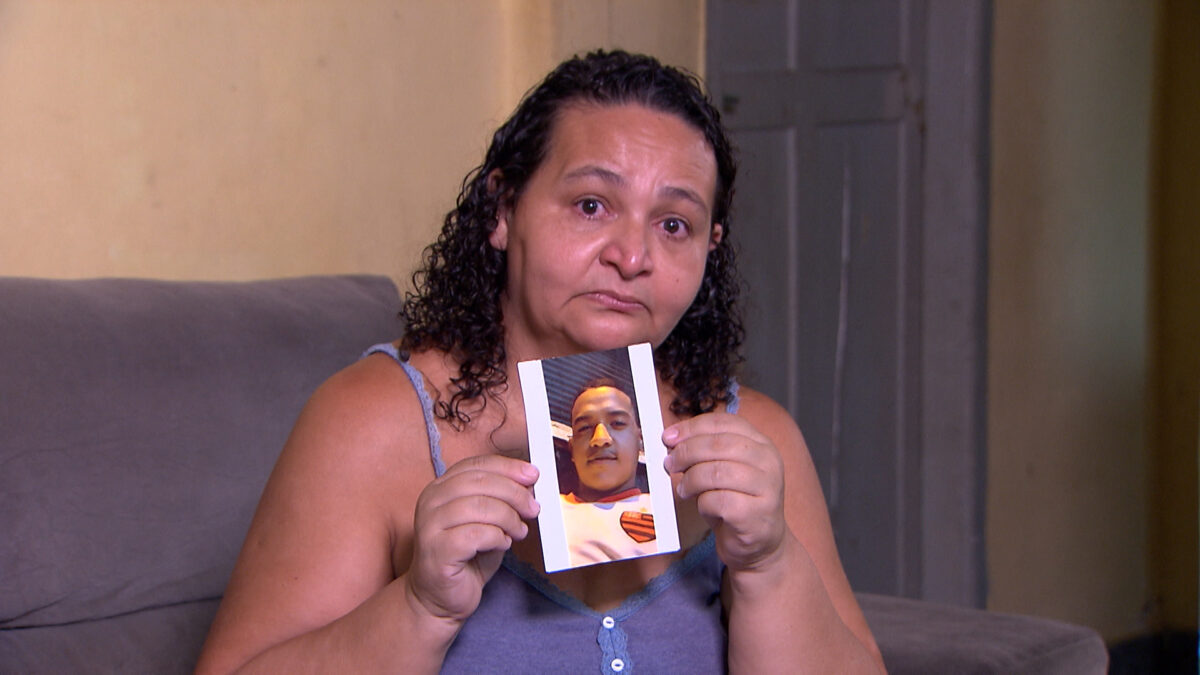 Sandra, sentada em um sofá com os olhos marejos e segurando em suas mãos uma foto do filho. Trecho da entrevista para o Repórter Record Investigação