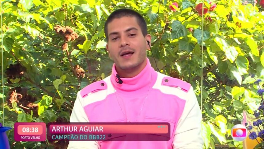 Arthur com uma roupa rosa com branco sentado numa mesa posta de café da manhã.