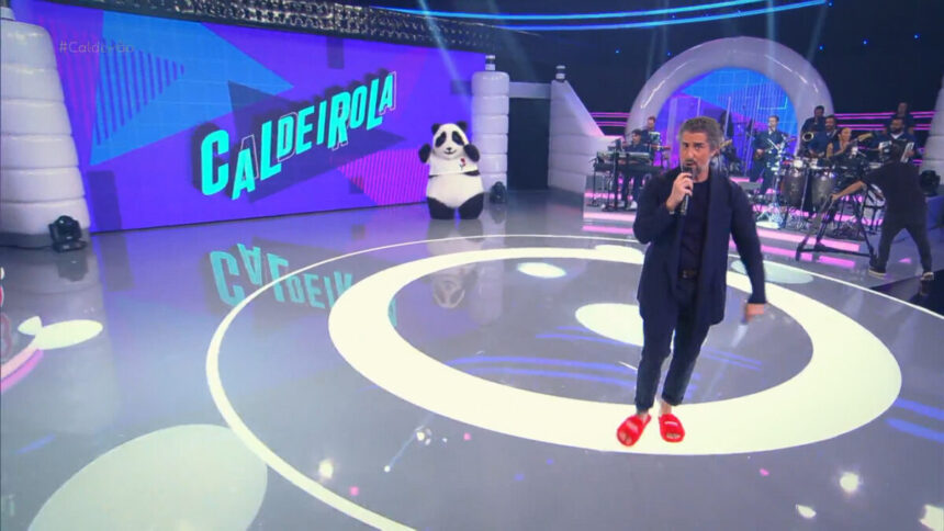 Marcos Mion no centro do palco do Caldeirão usando terno e pantufas laranjas