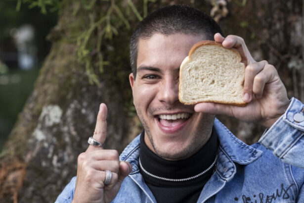 Arthur Aguiar apontando o dedo indicador para cima, com um pão de forma na mão, cobrindo um de seus olhos