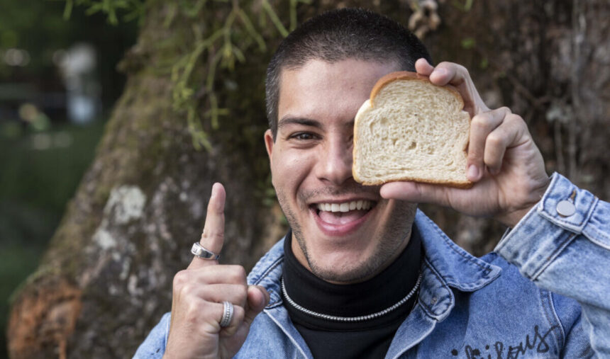 Arthur Aguiar apontando o dedo indicador para cima, com um pão de forma na mão, cobrindo um de seus olhos