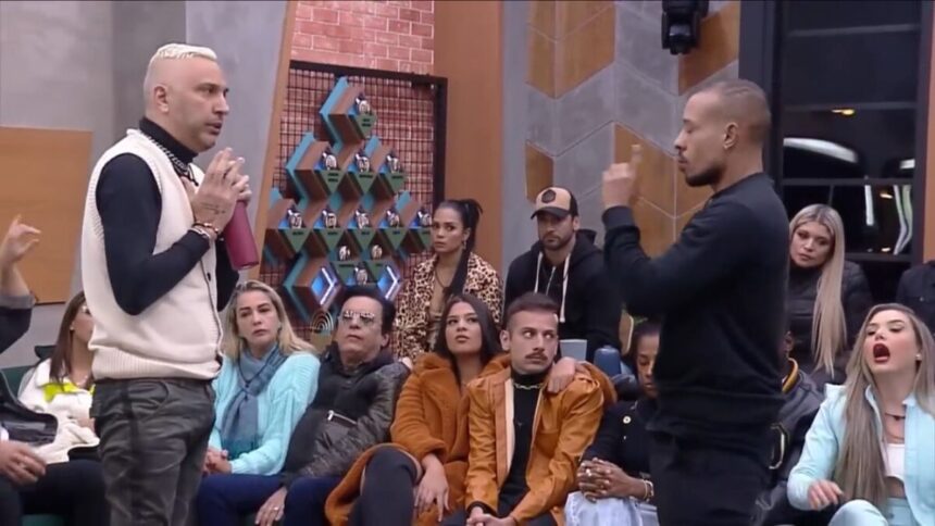 Rogério de roupa preta com branca de frente para Mussunzinho vestido de roupa preta. Ao redor deles estão os participantes do Power Couple Brasil 6.