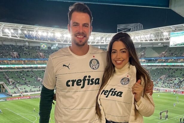 Gustavo e Laís abraçados, com a camisa do Palmeiras, tendo a visão do estádio do clube ao fundo