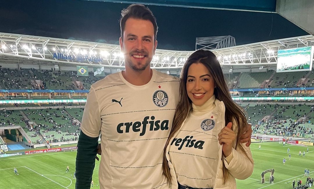 Gustavo e Laís abraçados, com a camisa do Palmeiras, tendo a visão do estádio do clube ao fundo