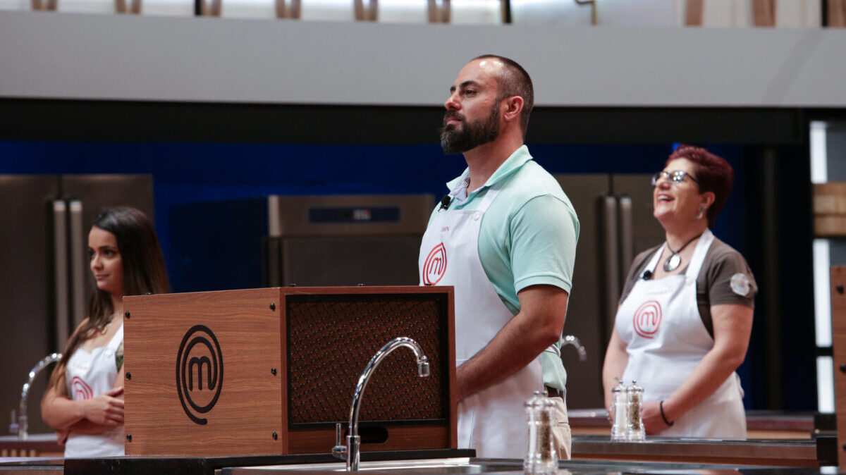 Trecho da estreia do MasterChef Brasil, com o desafio da Caixa Misteriosa na frente da imagem, com três participantes ao fundo