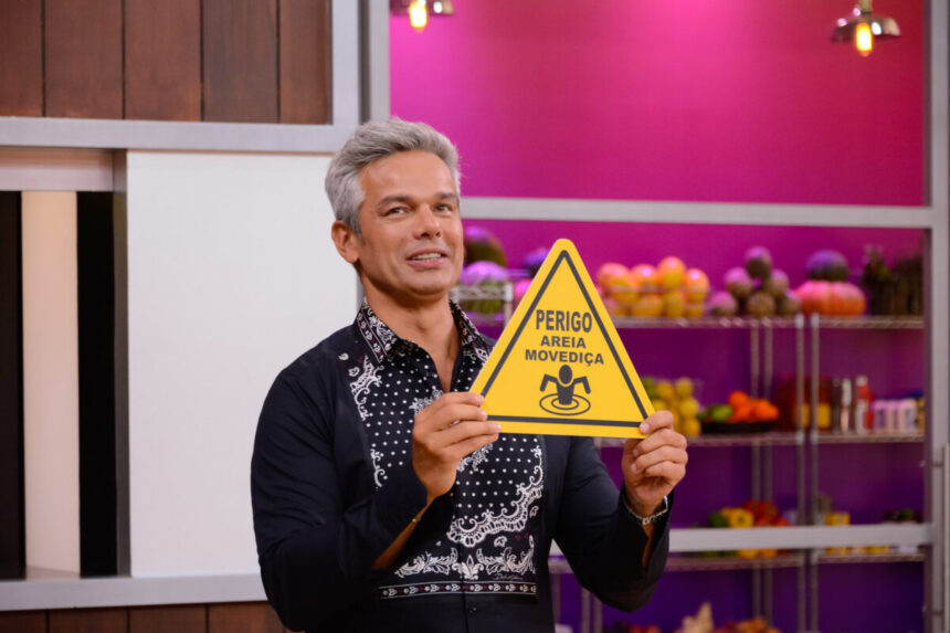 Otaviano Costa em trecho do terceiro episódio do reality Cozinhe se Puder. Ele segura uma placa escrita "Perigo: Areia Movediça"